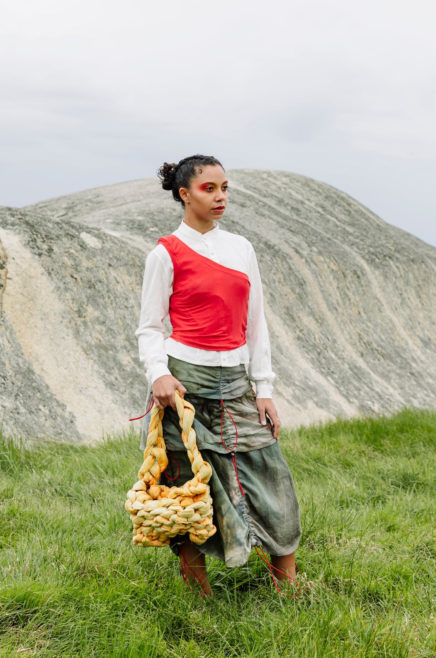 A woman stands in a grassy field with large rock formations in the background, holding a chunky knitted yellow and brown shoulder bag. She is wearing a very pale green long-sleeve top with a red asymmetrical tank and a dark green tiered skirt.