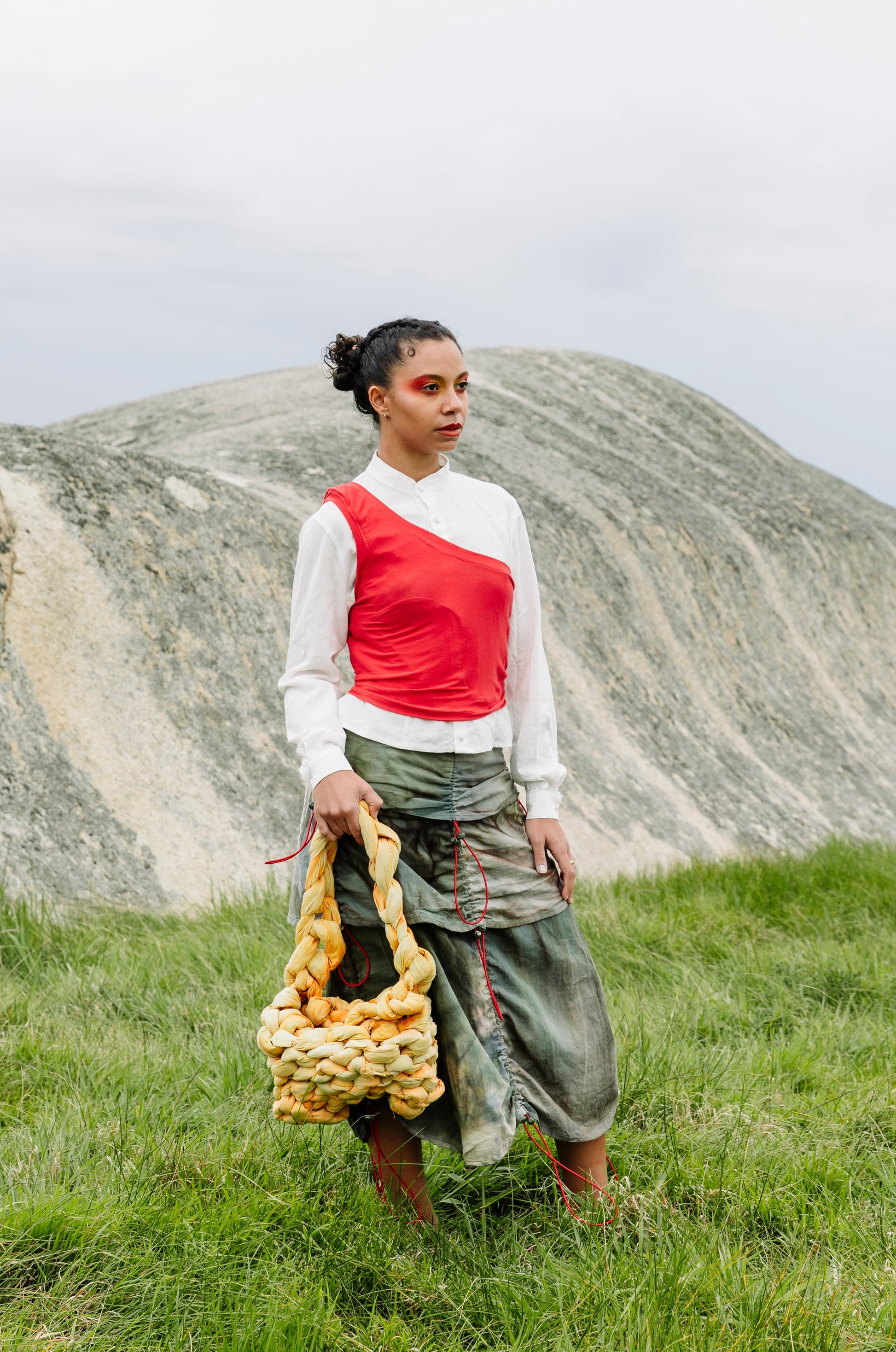 A woman stands in a grassy field with large rock formations in the background, holding a chunky knitted yellow and brown shoulder bag. She is wearing a very pale green long-sleeve top with a red asymmetrical tank and a dark green tiered skirt.